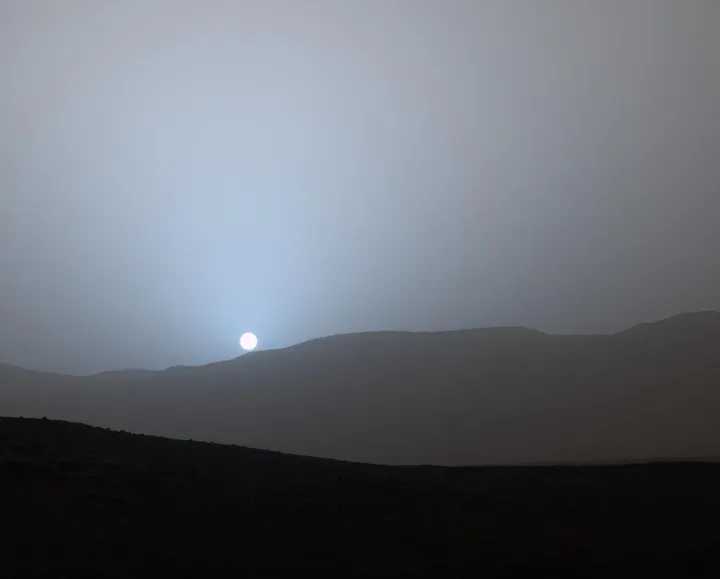 Tramonto nel cratere Gale su Marte: il Sole piccolo e bianco in un alone azzurro sopra le colline scure.